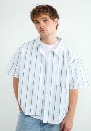 Młody mężczyzna z kręconymi włosami, ubrany w białą koszulę z krótkim rękawem z cienkimi pionowymi paskami na białym t-shircie, pozujący na jednolitym tle.