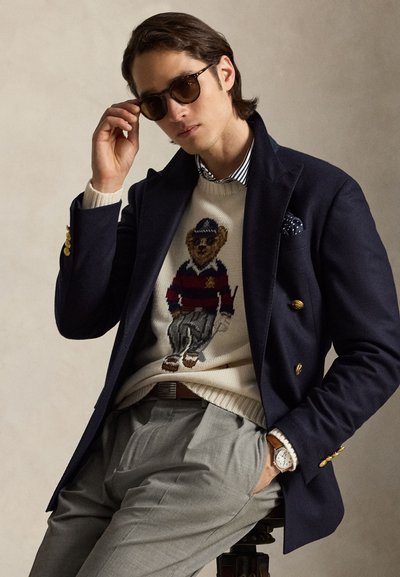 Homme portant des lunettes de soleil, un blazer bleu marine avec des boutons dorés, un pull à motif ours, un pantalon gris et une montre-bracelet, assis sur un tabouret.