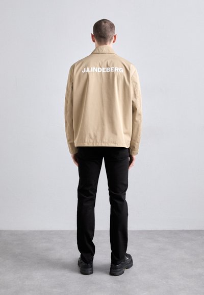Khakifarbene Bomberjacke mit "J.LINDEBERG"-Logo auf dem Rücken, kombiniert mit schwarzen Hosen und schwarzem Schuhwerk, von hinten angezeigt.