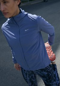 Violetti urheilu takki hupulla, kontrastoivalla vetoketjulla ja Nike-logoilla. Mustat kuviolliset legginsit ja punaruskeat lenkkarit näkyvät.