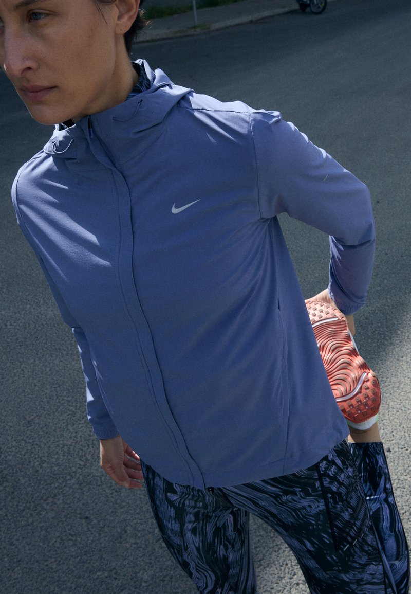 Violetti urheilu takki hupulla, kontrastoivalla vetoketjulla ja Nike-logoilla. Mustat kuviolliset legginsit ja punaruskeat lenkkarit näkyvät.
