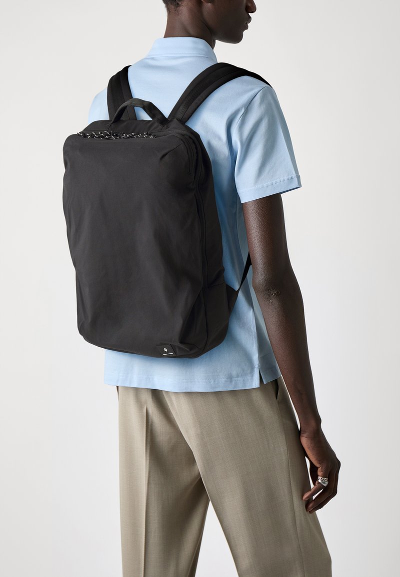 Personne portant une chemise polo bleu clair et un pantalon beige, portant un grand sac à dos noir avec des bretelles rembourrées sur le dos.