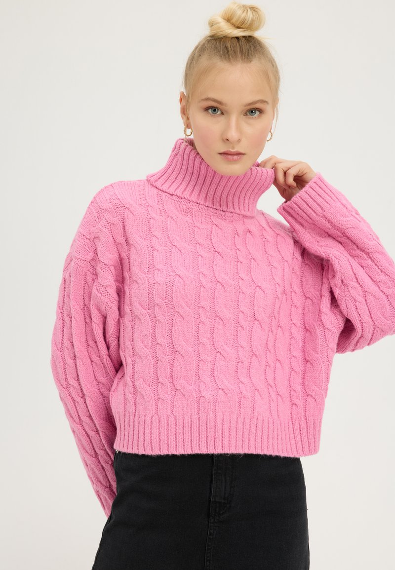 Pull en tricot rose avec un col montant côtelé haut et des manches oversized, présentant un motif texturé, un ourlet court et des poignets côtelés.
