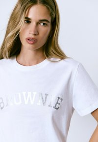 Camiseta blanca de algodón con la palabra "BROWNIE" en texto de purpurina plateada. Mangas cortas, cuello redondo, textura suave.