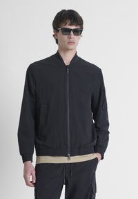Chaqueta bomber negra con cierre de cremallera en la parte delantera, puños y dobladillo acanalados. Presenta un bolsillo en la manga izquierda y está hecha de un tejido suave y ligero.