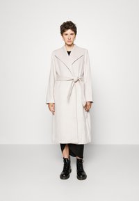 Abrigo largo beige con cinturón, solapas anchas y mangas largas, llevado sobre un vestido negro, combinado con botas negras de cordones.