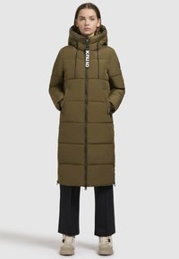 Olivovo zelená hrubá bunda s kapucňou, so zipsom na prednej strane, dvoma bočnými vreckami a dlhým strihom. V kombinácii s čiernymi nohavicami a béžovými topánkami.