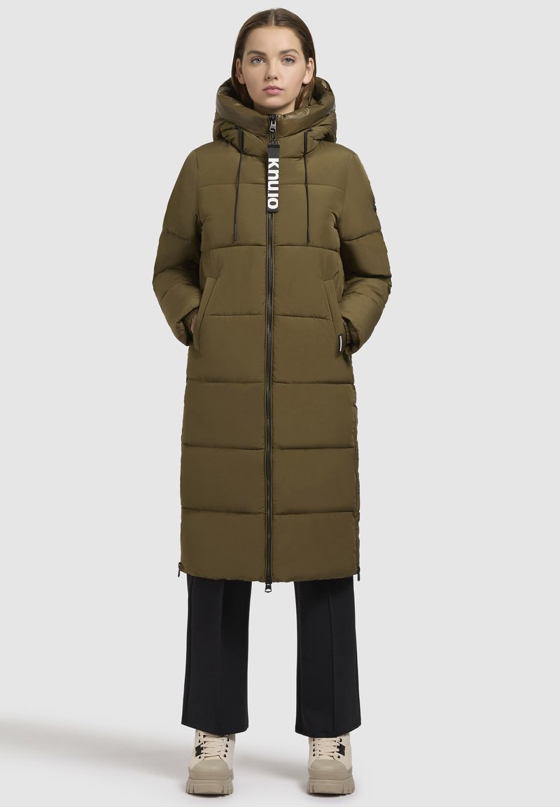 Olivovo zelená hrubá bunda s kapucňou, so zipsom na prednej strane, dvoma bočnými vreckami a dlhým strihom. V kombinácii s čiernymi nohavicami a béžovými topánkami.