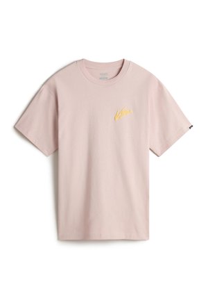 Personne portant un t-shirt rose clair à manches courtes avec le logo Vans jaune sur la poitrine et un pantalon beige clair, les mains dans les poches.