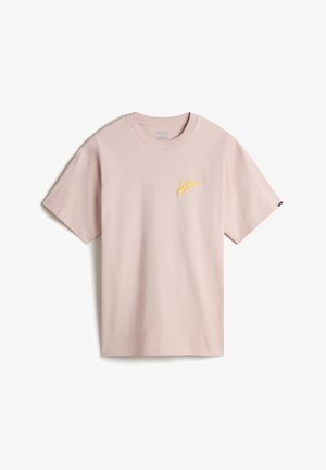Person trägt ein hellrosa kurzärmeliges T-Shirt mit gelbem Vans-Logo auf der Brust und hellbeige Hose, die Hände in den Taschen.