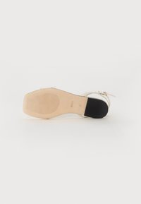 White leather heeled sandal with a narrow toe strap, adjustable ankle strap, and a textured beige rubber sole featuring a black accent.