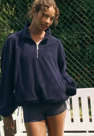 Marineblauer Fleecepullover mit einem Reißverschluss am Stehkragen und elastischem Saum. Verfügt über ein kleines Logo und voluminöse Ärmel, kombiniert mit dunklen Shorts.