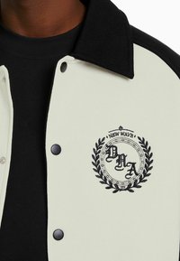 Vit och svart varsityjacka med knäppning fram, broderad cirkulär logotyp och strukturerad krage. Lagd över en svart skjorta.