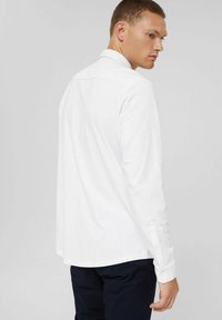 White long-sleeve shirt with a collar, straight cut, buttoned cuffs, and a smooth texture. Worn with dark pants, viewed from the back.