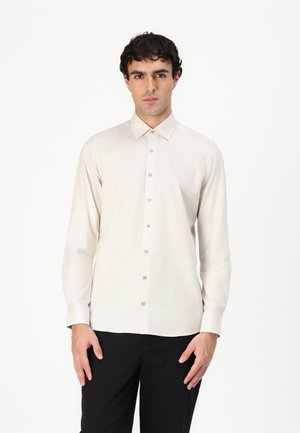 Camică bej deschis cu mânecă lungă, cu nasturi și guler, având o textură netedă, nasturi asortați și croială ajustată. Purtați cu pantaloni negri.