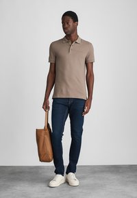 Hellbraunes Poloshirt mit einem dezenten Kragen, kombiniert mit dunkelblauen Jeans und weißen Sneakers. Das Model hält eine helle Ledertasche.