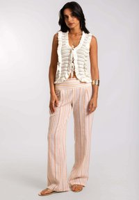 Chaleco de punto blanco con bordes rizados y lazo al frente, combinado con pantalones de pierna ancha a rayas claras en naranja y beige. Sandalias visibles.