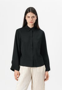 Svart långärmad skjorta med krage, texturerad tyg och en enda bröstficka. Bärs med ljusa, veckade byxor.