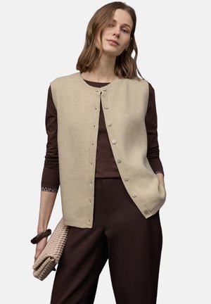 Beige ribstrikket vest med rund hals og knaplukning, båret over en brun top. Modellen holder en flettet clutchtaske og bærer et armbånd.