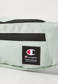 Sac banane en tissu vert clair avec une fermeture éclair noire. Présente un patch logo Champion et une surface texturée pour plus de durabilité.