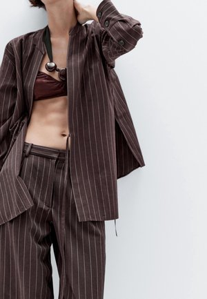 Femme portant un costume marron à fines rayures avec une veste courte et un pantalon, associé à un bralette bordeaux et un grand collier noir à perles, main derrière la nuque.