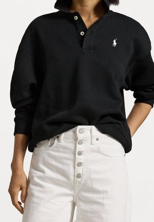 Personne portant un polo à manches longues noir avec un logo blanc brodé et un jean blanc taille haute à boutons apparents.
