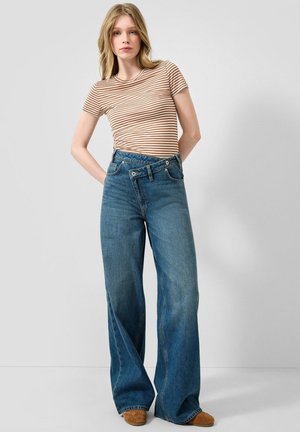 Junge Frau trägt ein braun-weiß gestreiftes Kurzarmtop mit weitem Bein blauen Jeans und braunen Schuhen, steht vor einem schlichten Hintergrund.