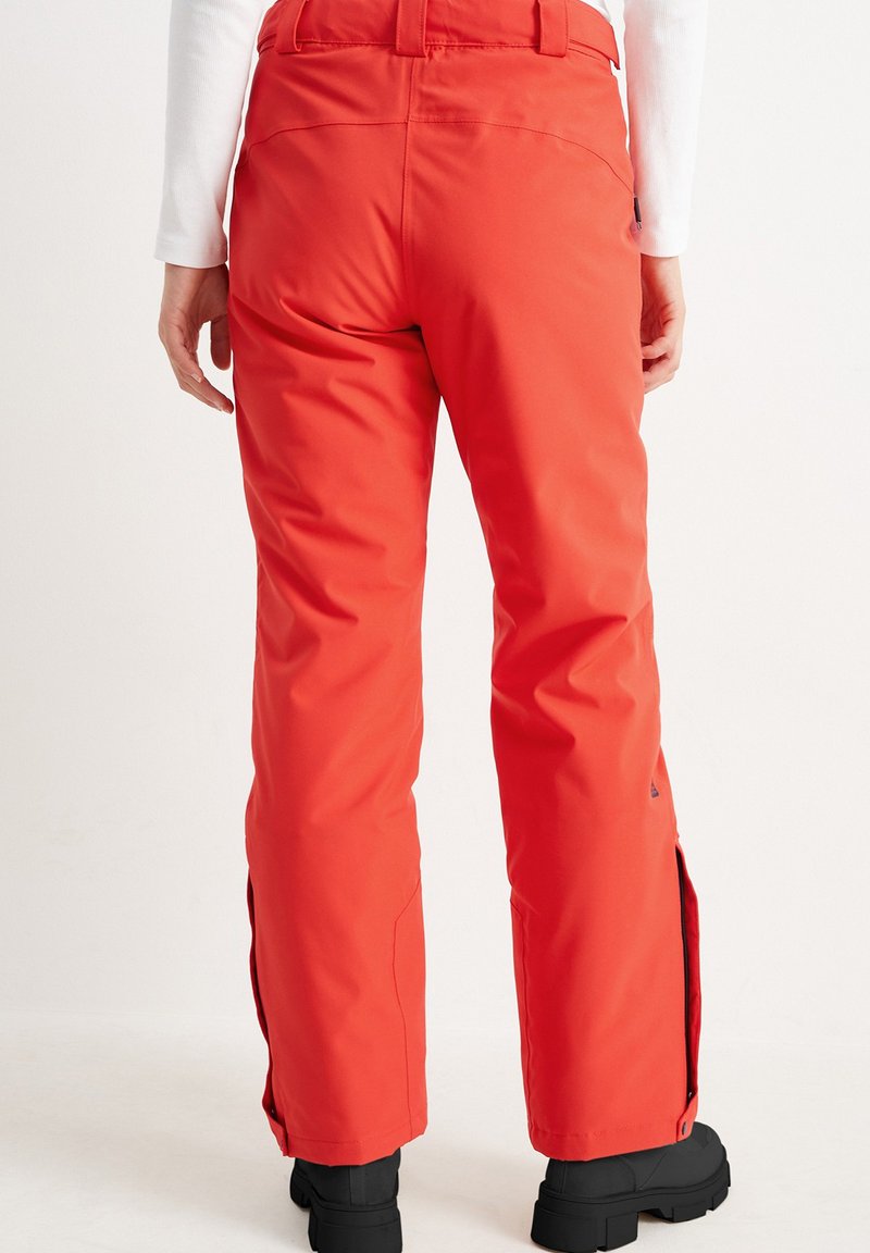 Person, der leuchtend rote, gerade geschnittene Hose und schwarze Stiefel trägt, steht vor einem schlichten weißen Hintergrund, von hinten betrachtet.