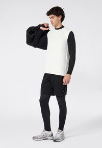 Giovane uomo che indossa leggings neri, pantaloni corti, una maglietta senza maniche bianca sopra una camicia a maniche lunghe nera, con una borsa da palestra nera a tracolla.