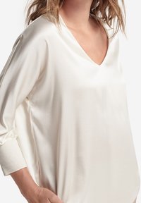 Žena v volné, lesklé krémové saténové blůze s dlouhými rukávy a výstřihem do V, ruka položená v kapse kalhot.