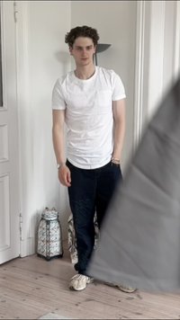 Hvit T-skjorte med korte ermer, mørkeblå jeans med mønster, beige joggesko, står i et lyst rom med lys tregulv og dekorative gjenstander.