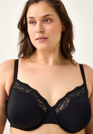 Kvinde med mellembrunt hår i mellem længde iført en sort bh med blonder og bøjle, der kigger let til siden mod en ensfarvet baggrund.