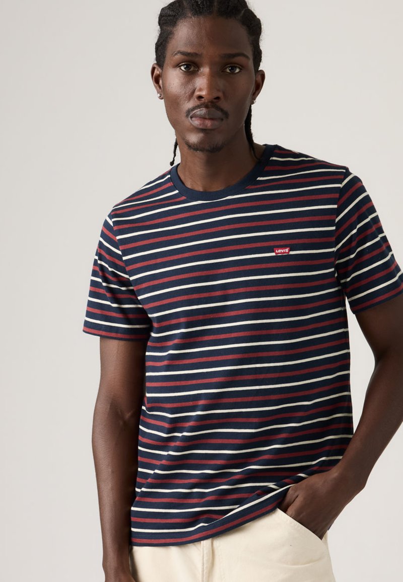 Mann mit geflochtenen Haaren, der ein marineblaues, rot-weiß gestreiftes Levi's-T-Shirt und beige Hosen trägt, eine Hand in der Tasche, neutraler Hintergrund.