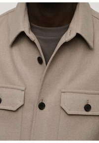 Beige wollen overhemdjasje met een kraag, twee borstzakken en zwarte knopen. De stof heeft een zachte textuur en een subtiele afwerking.