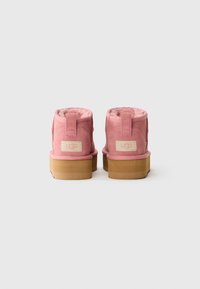 Une paire de bottes UGG roses avec doublure en polaire, semelles compensées brunes et patch de marque sur le talon, montrées de l'arrière sur un fond blanc.