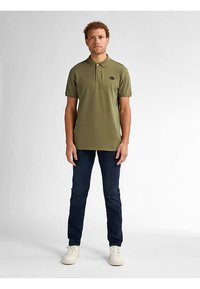 Tricou polo verde măsliniu cu mâneci scurte, guler și emblema neagră, asortat cu blugi bleumarin închiși și adidași albi.