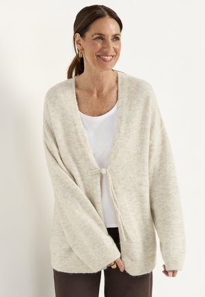 Vrouw met bruin haar naar achter gebonden, draagbaar gouden oorringen, een wit topje en een beige gebreide cardigan voorgebonden, glimlacht licht.