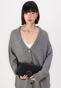 Cardigan gris de punto con un escote en V pronunciado y cierre de botones, llevado sobre una blusa blanca. Sosteniendo un bolso de mano de cuero negro con un detalle de hebilla metálica.