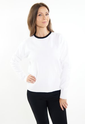 Weißer, gestrickter Pullover mit rundem, schwarzem Halsausschnitt, langen Ärmeln, gerippten Bündchen und Saum. Tragend über schwarzen Hosen.