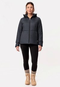 Zwarte geïsoleerde jas met capuchon, ritssluiting en gestikt ontwerp. Draagt over zwarte legging en tankleurige laarzen met gestructureerde accenten.