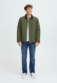 Giacca imbottita verde con colletto in velluto a coste marrone, abbinata a jeans blu e sneaker bianche. La giacca presenta bottoni a pressione e una vestibilità morbida.