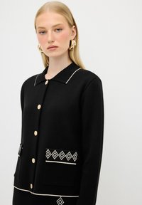Svart kardigan med guldknappar, vit kantad krage, strukturerad tyg, två framfickor med geometriska mönster.
