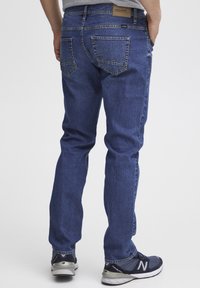 Jeans de mezclilla azul oscuro con corte recto, costuras visibles y dos bolsillos traseros con bordado en forma de montaña. Llevados con zapatillas.