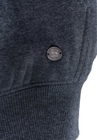 Jersey gris oscuro con puños acanalados y un botón de metal con detalle del logo. La tela parece suave y tiene una textura lisa.