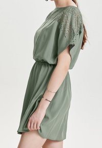 Vestido corto verde de tejido ligero con detalles de encaje en los hombros, cintura elástica y falda fluida. Textura suave y corte holgado.