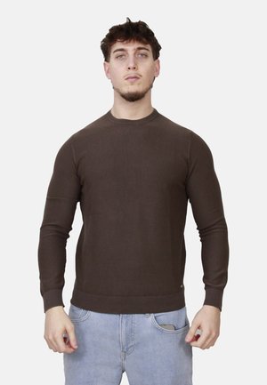 Giovane uomo in piedi che indossa un maglione marrone aderente a maniche lunghe e jeans azzurri con uno smartphone nella tasca destra.