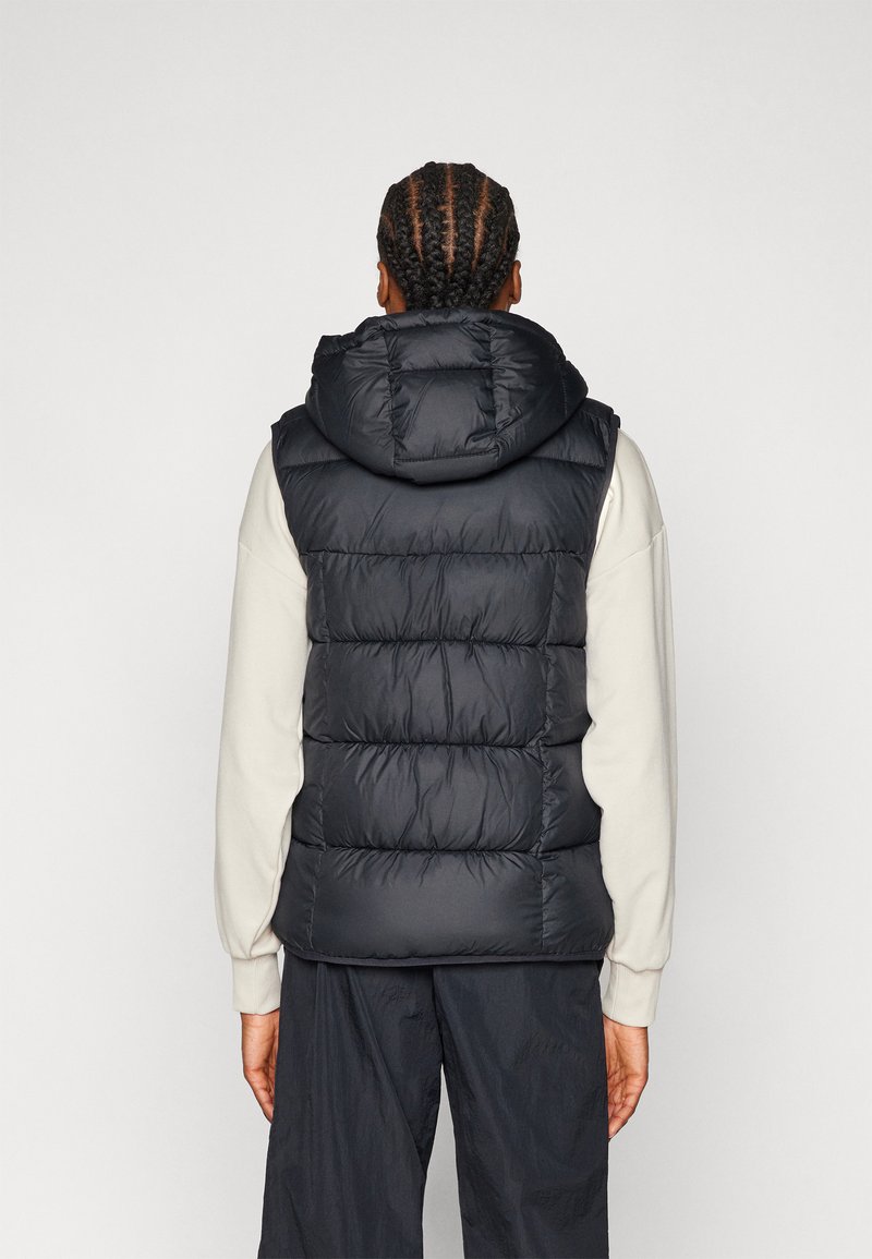 Czarna kamizelka puchowa z kapturem, z pikowanym wzorem i grubym wypełnieniem. Noszona na jasnej długim rękawem bluzce.