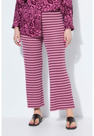 Person iført lyserøde mønstrede bukser, en lilla blomsterprintet top, sorte sandaler og flere ringe på begge hænder, der står op ad en hvid væg.