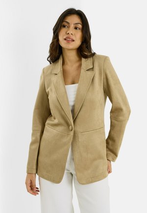 Femme portant un blazer en daim beige à un bouton par-dessus un haut blanc et un pantalon blanc, regardant légèrement sur le côté avec une pose détendue.
