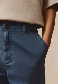 Pantalones de algodón azules con textura suave, corte ajustado y bolsillos laterales, asegurados con un botón marrón oscuro en la cintura.